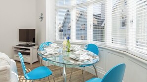 A modern dining area with a glass table, blue chairs, and a TV on a white cabinet. - Charming Apartment with Garden & Parking (Dartmouth)
