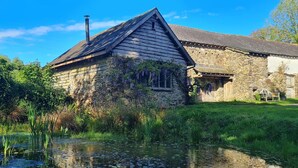 A rustic stone building with a sloped roof, a chimney, and a pond in the foreground. - Escape to an Idyllic Countryside Couple's Retreat (Totnes)