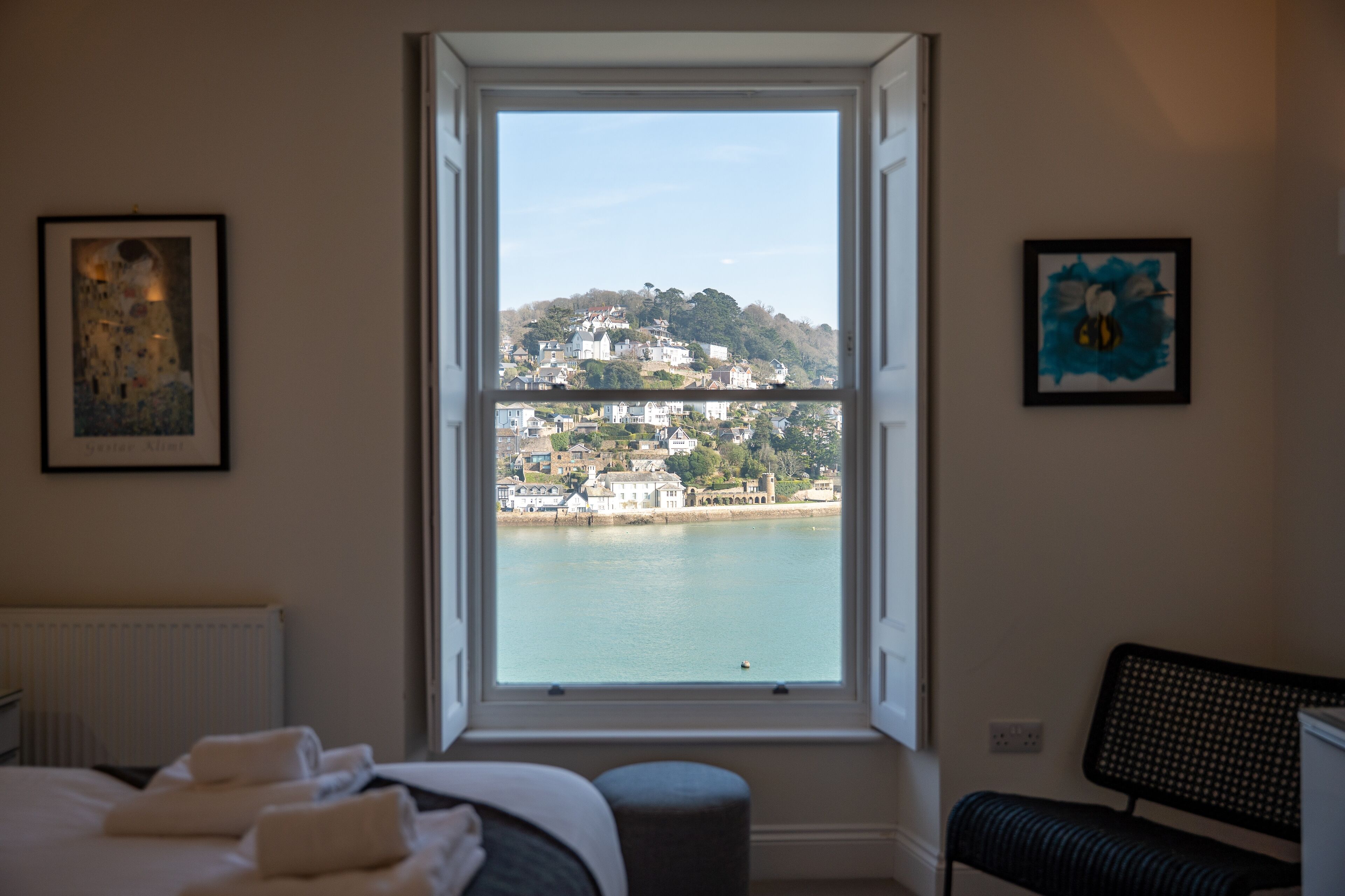 A bedroom with a large window overlooking a coastal town, a bed with neatly folded towels, and two framed artworks on the walls.