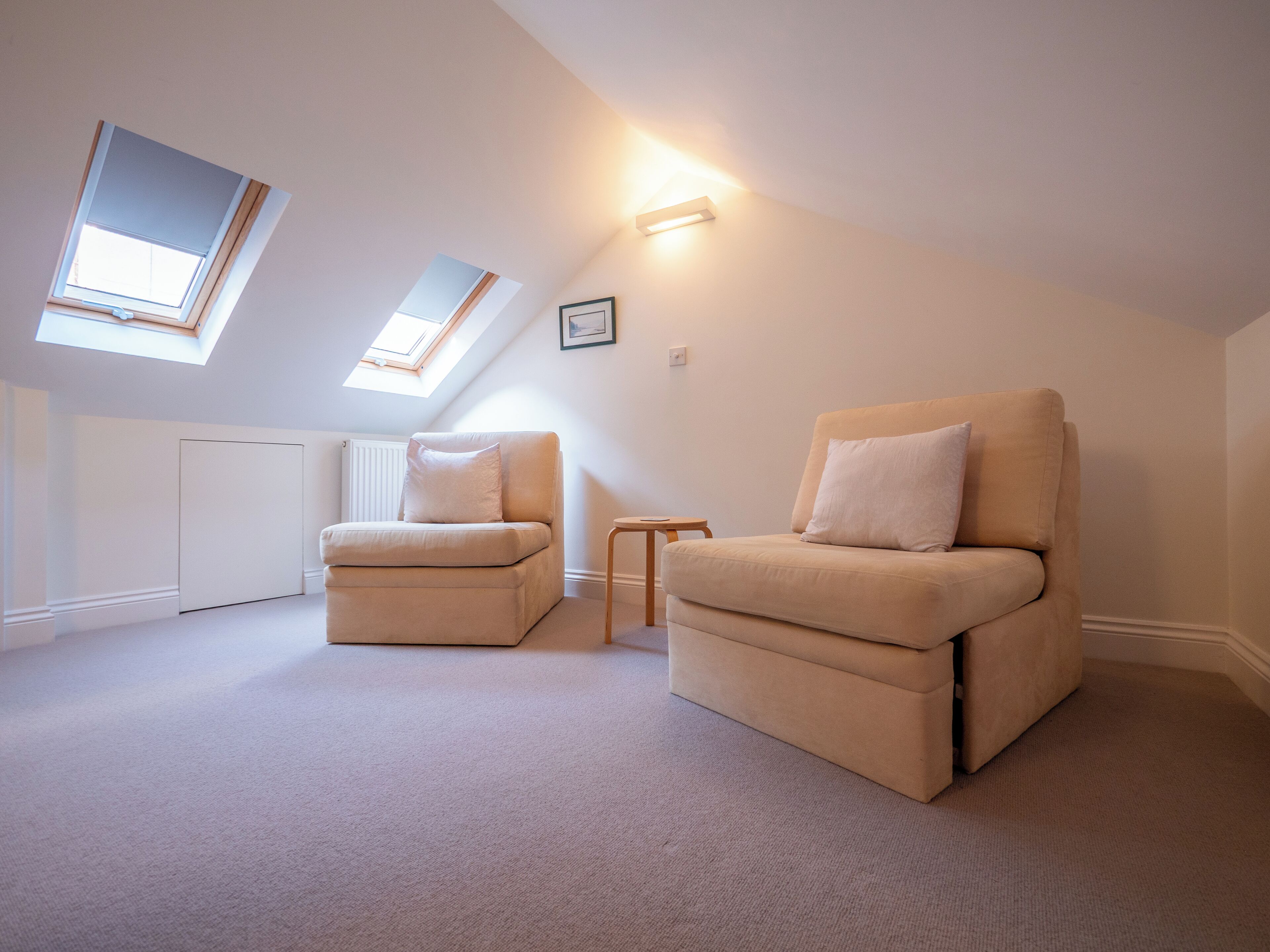 A spacious attic room with two beige armchairs, a small wooden side table, and skylights providing natural light.