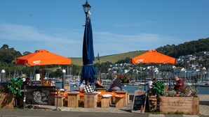 Un bar sul lungomare con tavoli di legno e ombrelloni arancioni, affacciato su un porto turistico e una cittadina in collina.