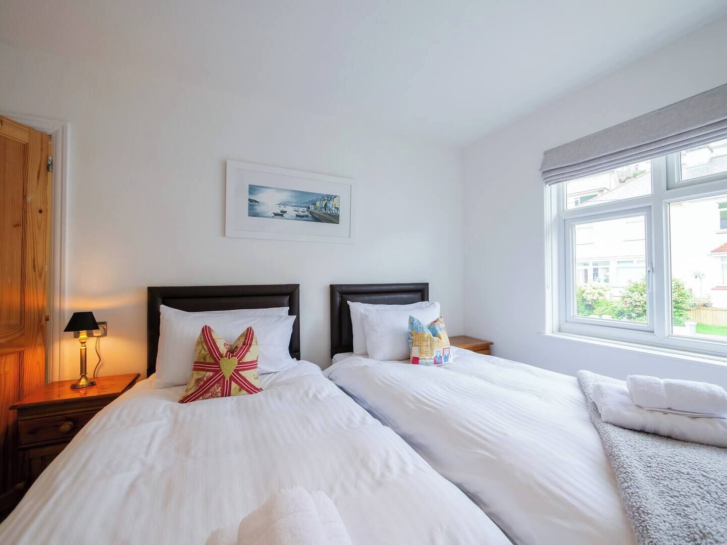 A double room with two beds, a nightstand with a lamp, a framed picture on the wall, and a window with a view of the outside.