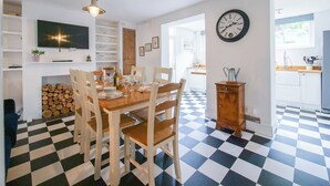 Zona pranzo con pavimento a scacchi, tavolo in legno e sedie. C'è una televisione a parete, una sveglia e un pensile con utensili da cucina.
