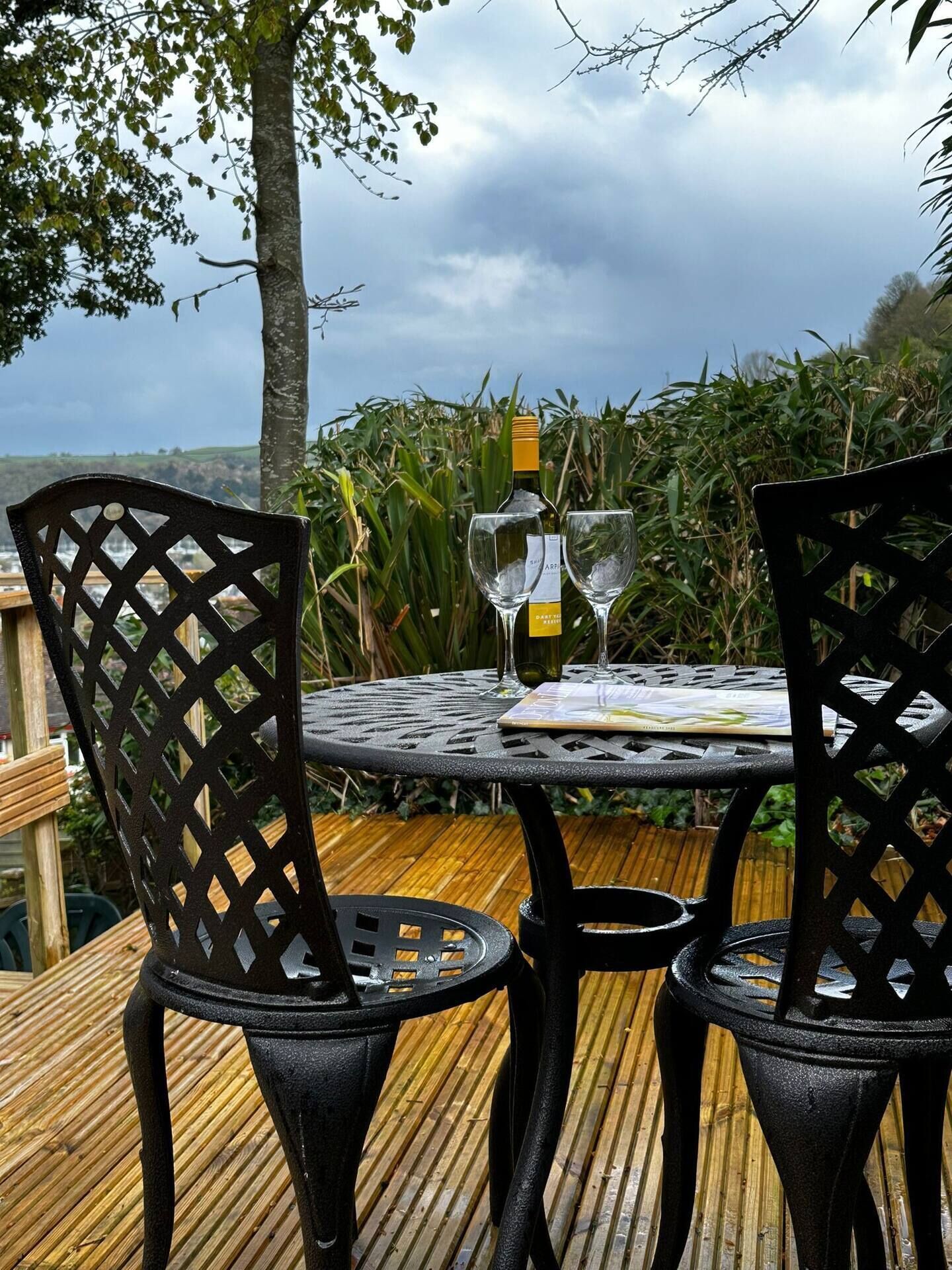 Ein Tisch mit zwei Stühlen, einer Weinflasche und einem Glas auf einer Terrasse mit Blick auf ein Feld und Bäume.