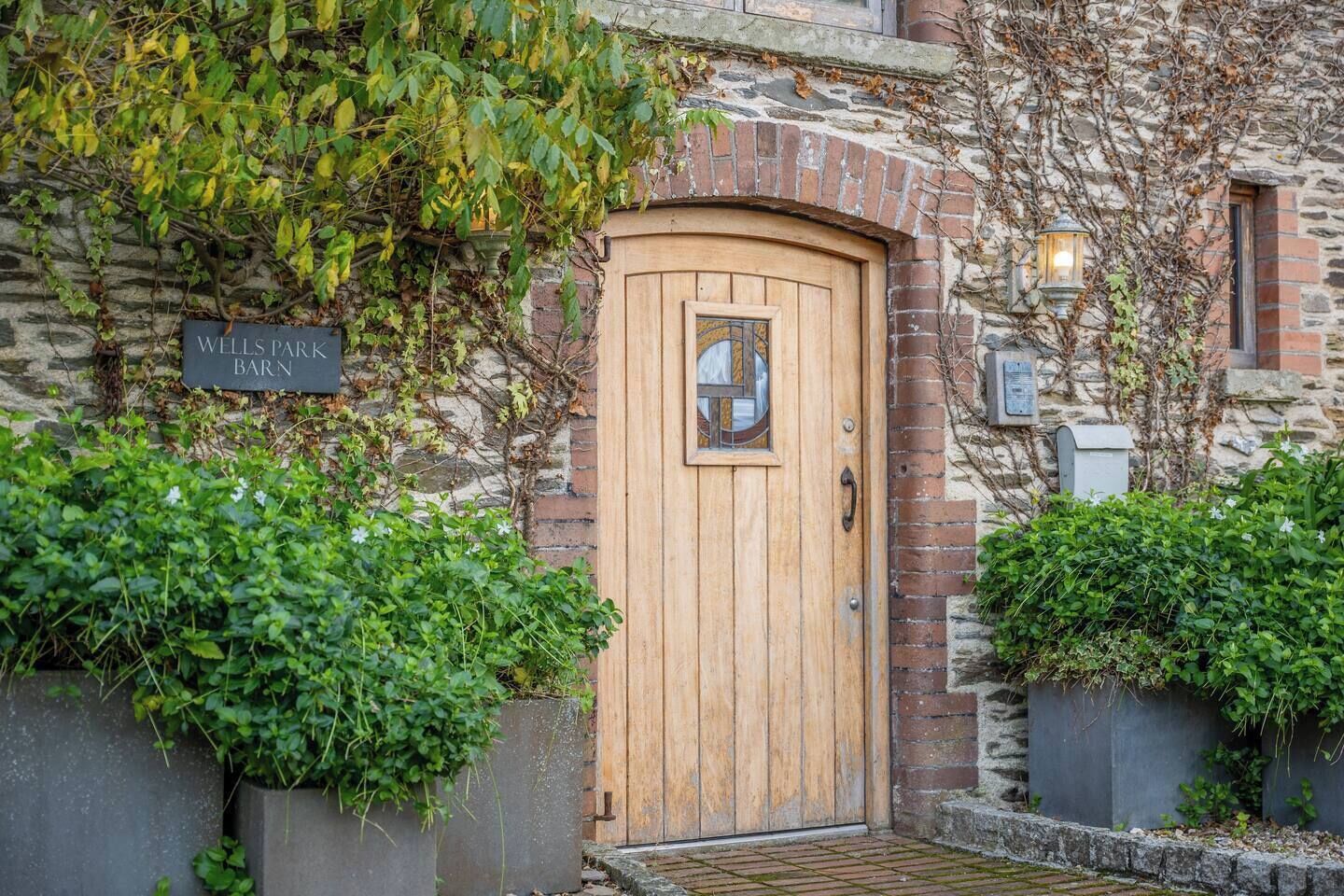 Ein rustikales Steinhaus mit einer Holztür, einem Schild mit der Aufschrift 'WELLS PARK BARN' und einem Laternen am rechten Rand.