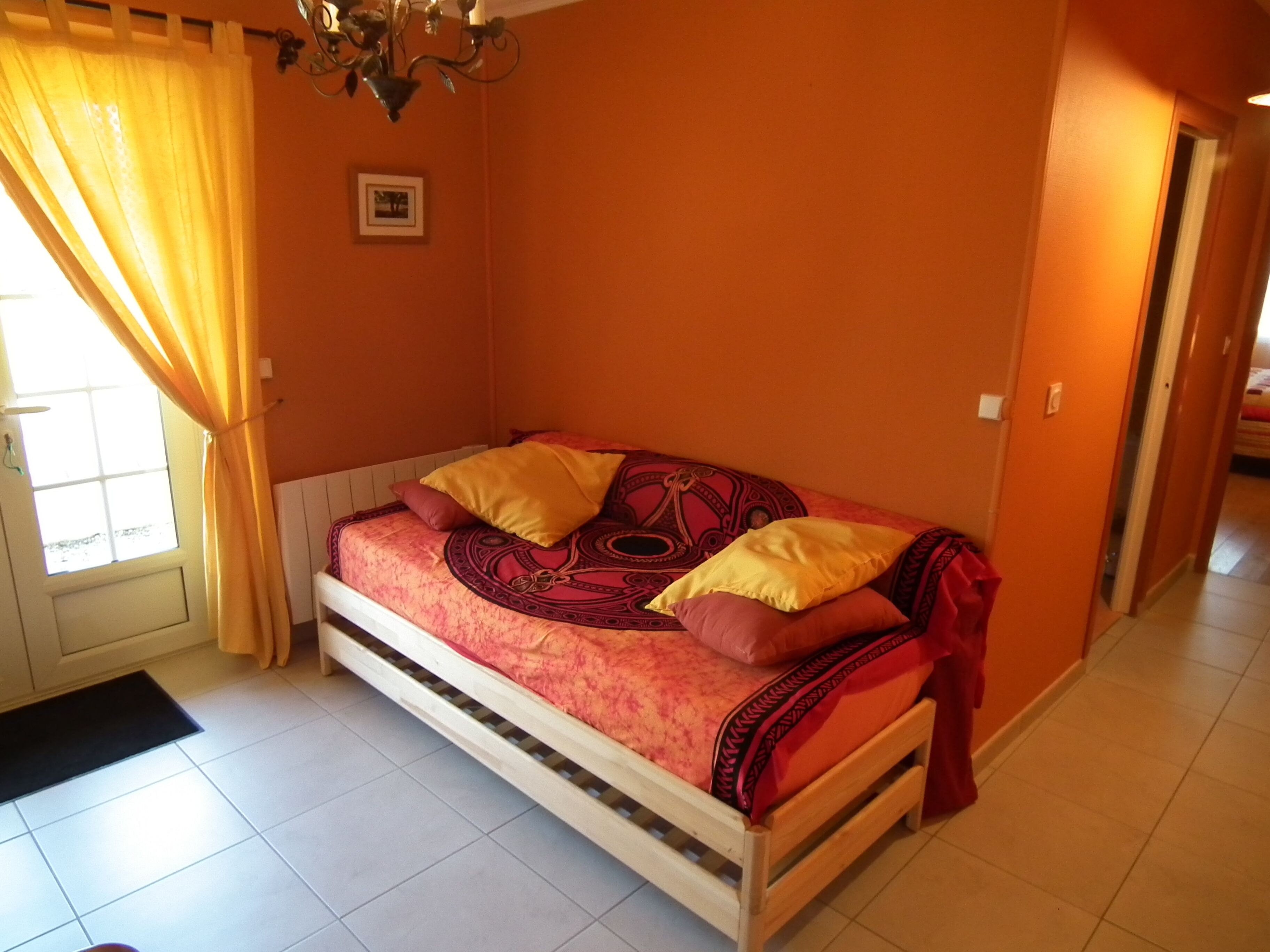 Una cama individual en una habitación con paredes anaranjadas, una ventana con cortinas amarillas y un cuadro en la pared.
