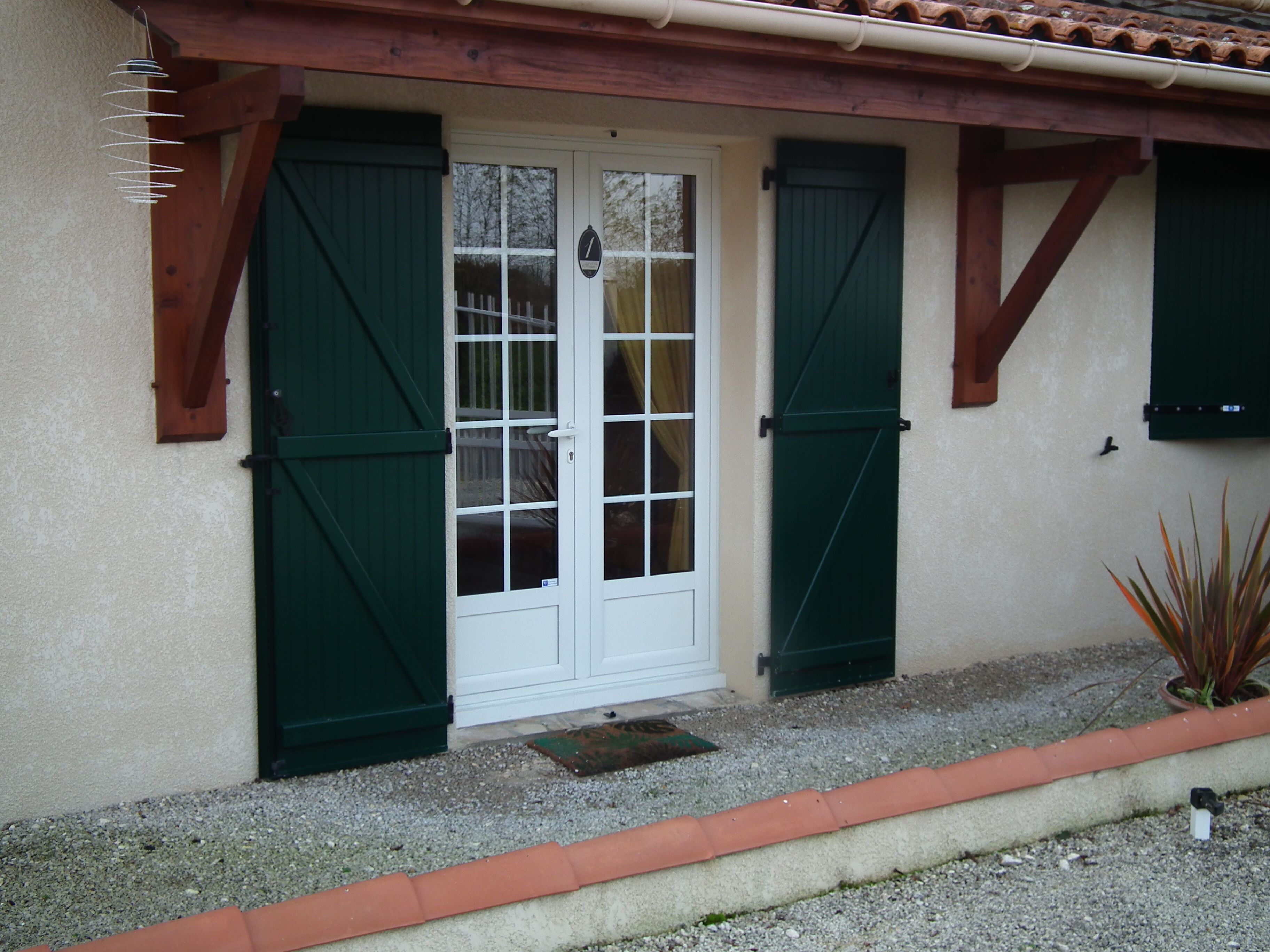 Una casa con puerta blanca, contraventanas verdes y un pequeño jardín con grava y una planta en maceta.
