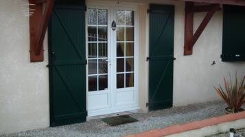 Una casa con puerta blanca, contraventanas verdes y un pequeño jardín con grava y una planta en maceta.
