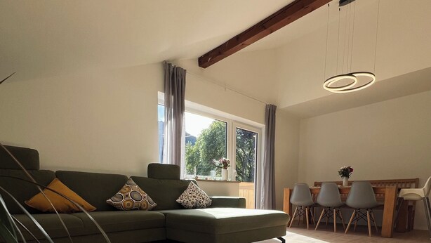A modern living room with a sectional sofa, a dining area with a wooden table and chairs, and large windows with curtains.