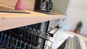 A kitchen with a dishwasher, a coffee maker, and a pink vase with flowers on a wooden shelf.