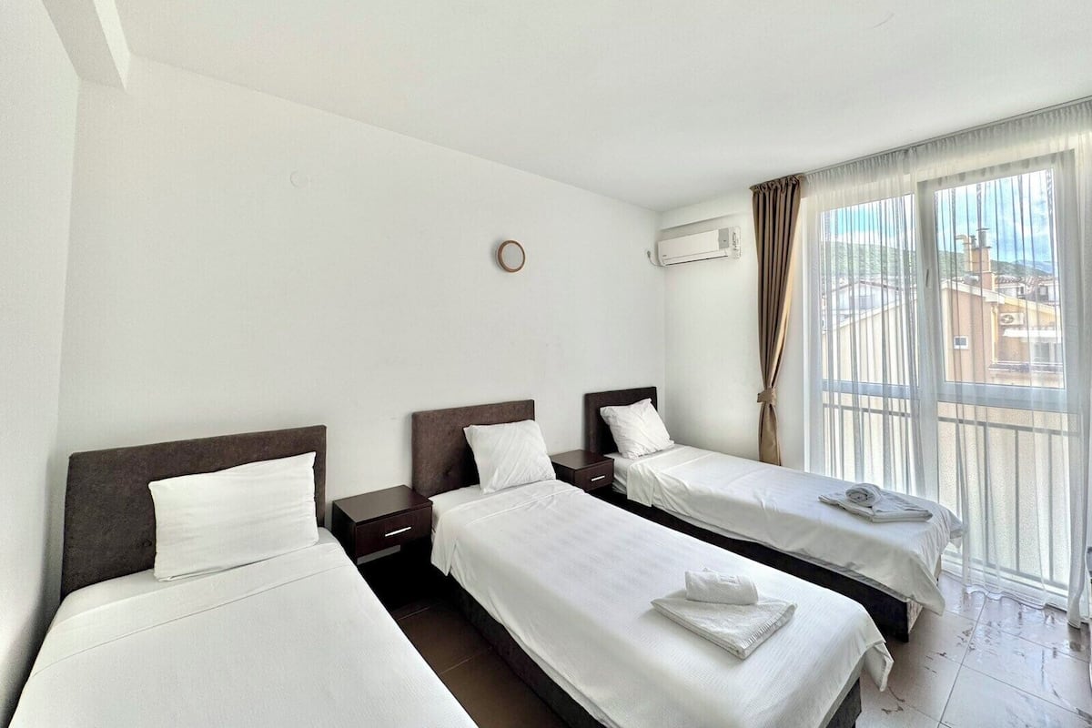 Una habitación de hotel con tres camas individuales, una mesita de noche pequeña con lámpara, una ventana con cortinas y un equipo de aire acondicionado en la pared.