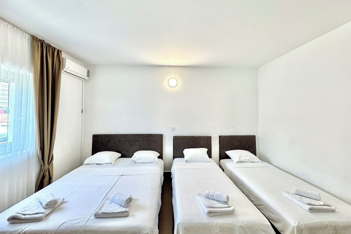 Habitación de hotel con dos camas individuales, cada una con una almohada y una toalla enrollada. Hay una ventana con cortinas, un equipo de aire acondicionado y un aplique de pared.