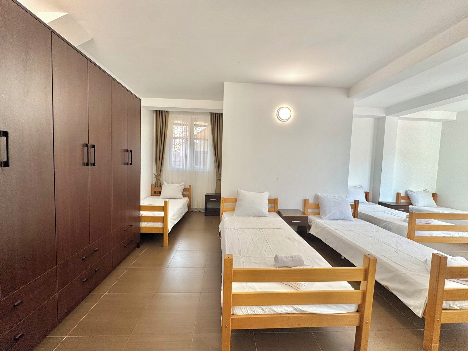 A dormitory room with four single beds, a wardrobe, and a small window with curtains.