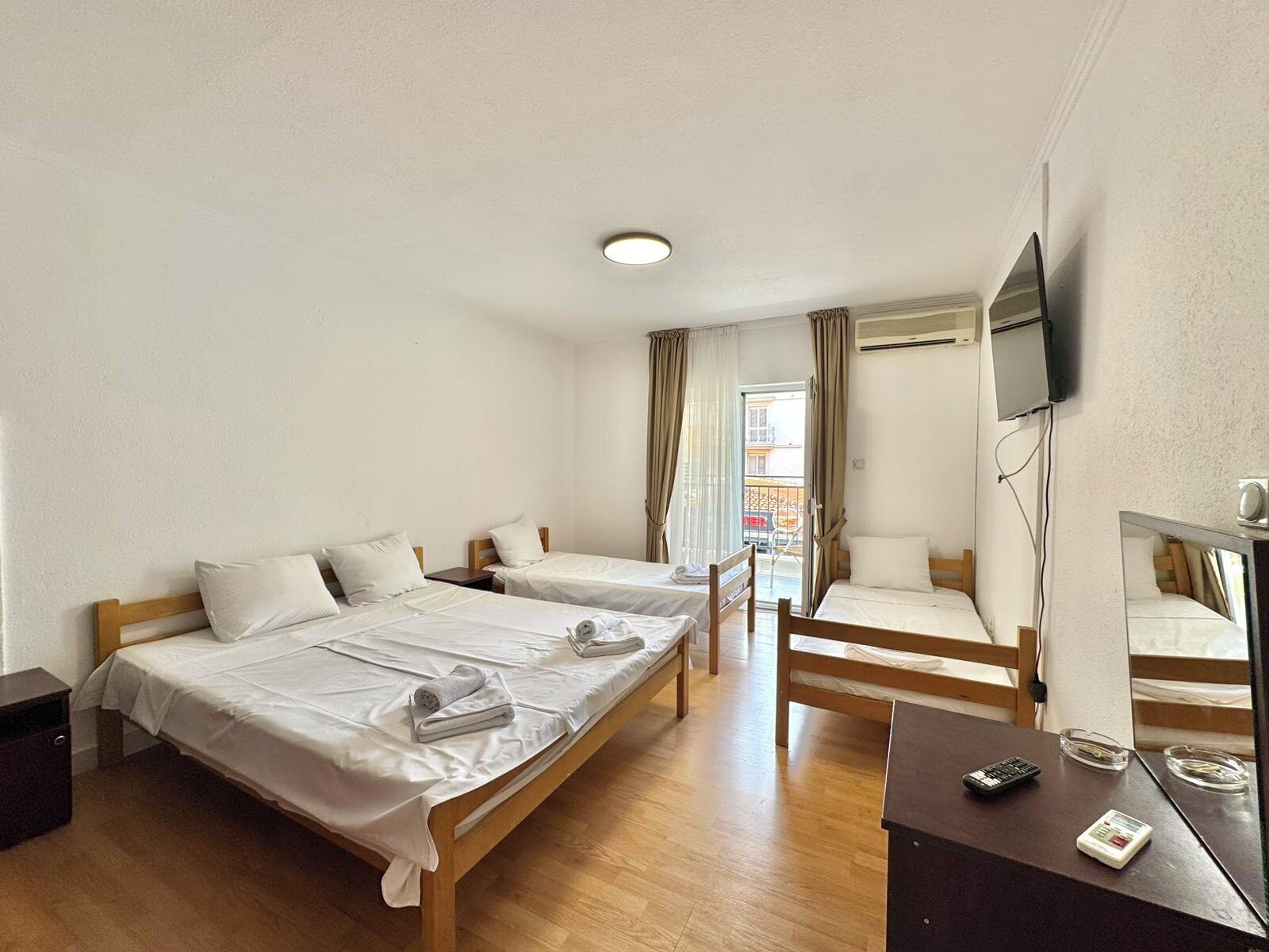 A hotel room with two single beds, a wooden desk, a TV mounted on the wall, and a balcony with a view.