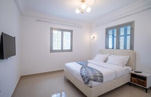 A bedroom with a bed, a TV, a nightstand, and two windows. - Dream Garden Djerba (Mezraia)