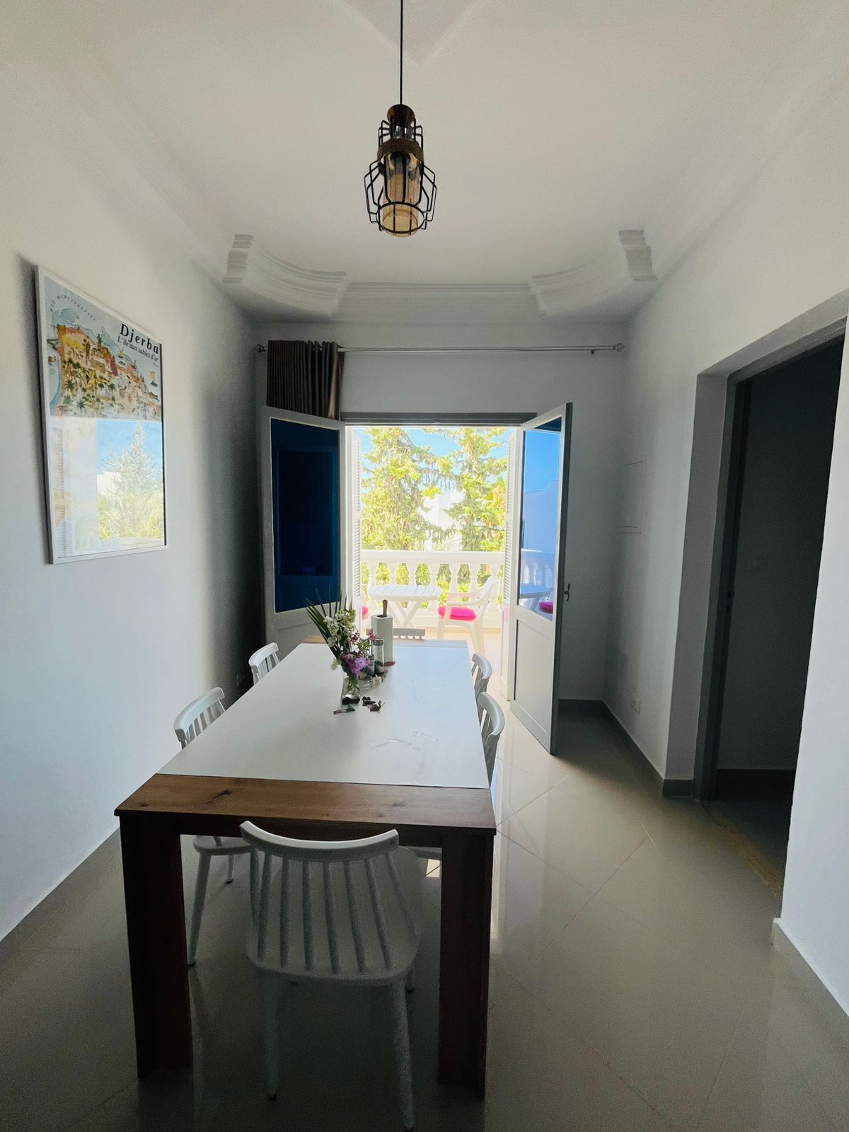Un comedor con mesa y sillas blancas, una lámpara colgante y vistas a un área exterior con cerca blanca y follaje.