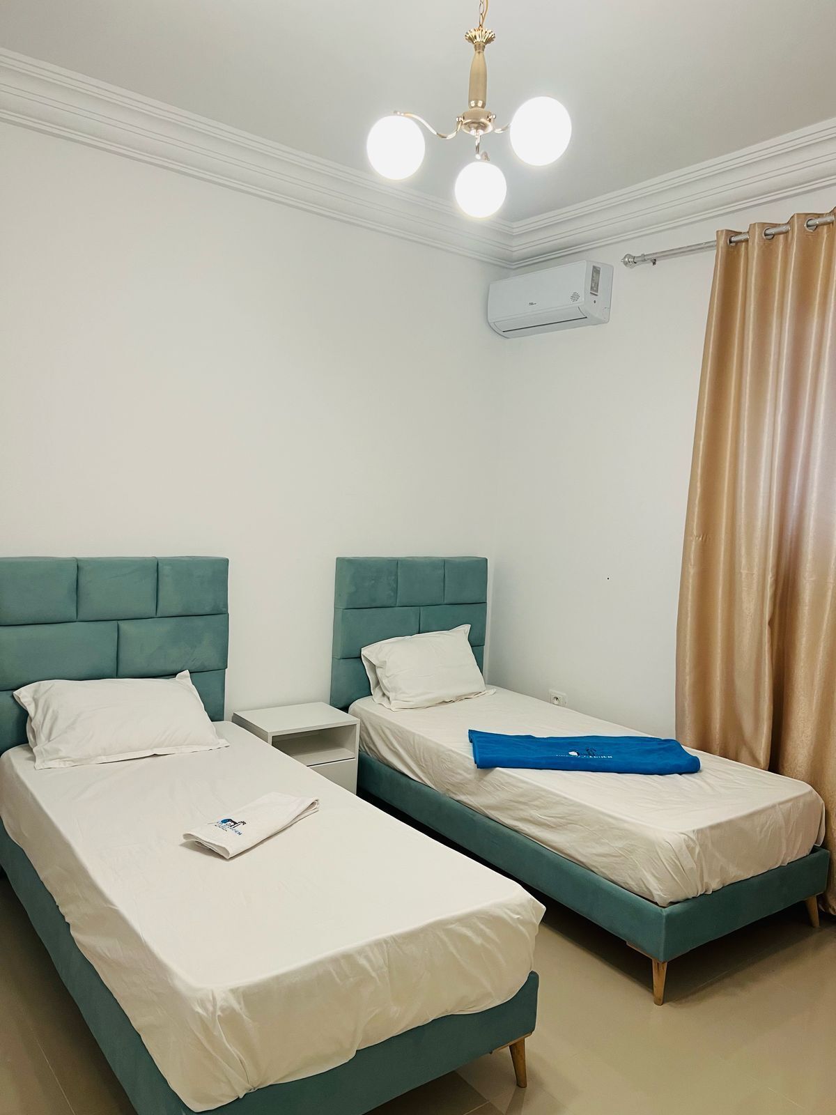 A hotel room with two single beds, a nightstand, a ceiling light fixture, an air conditioning unit, and a curtain.