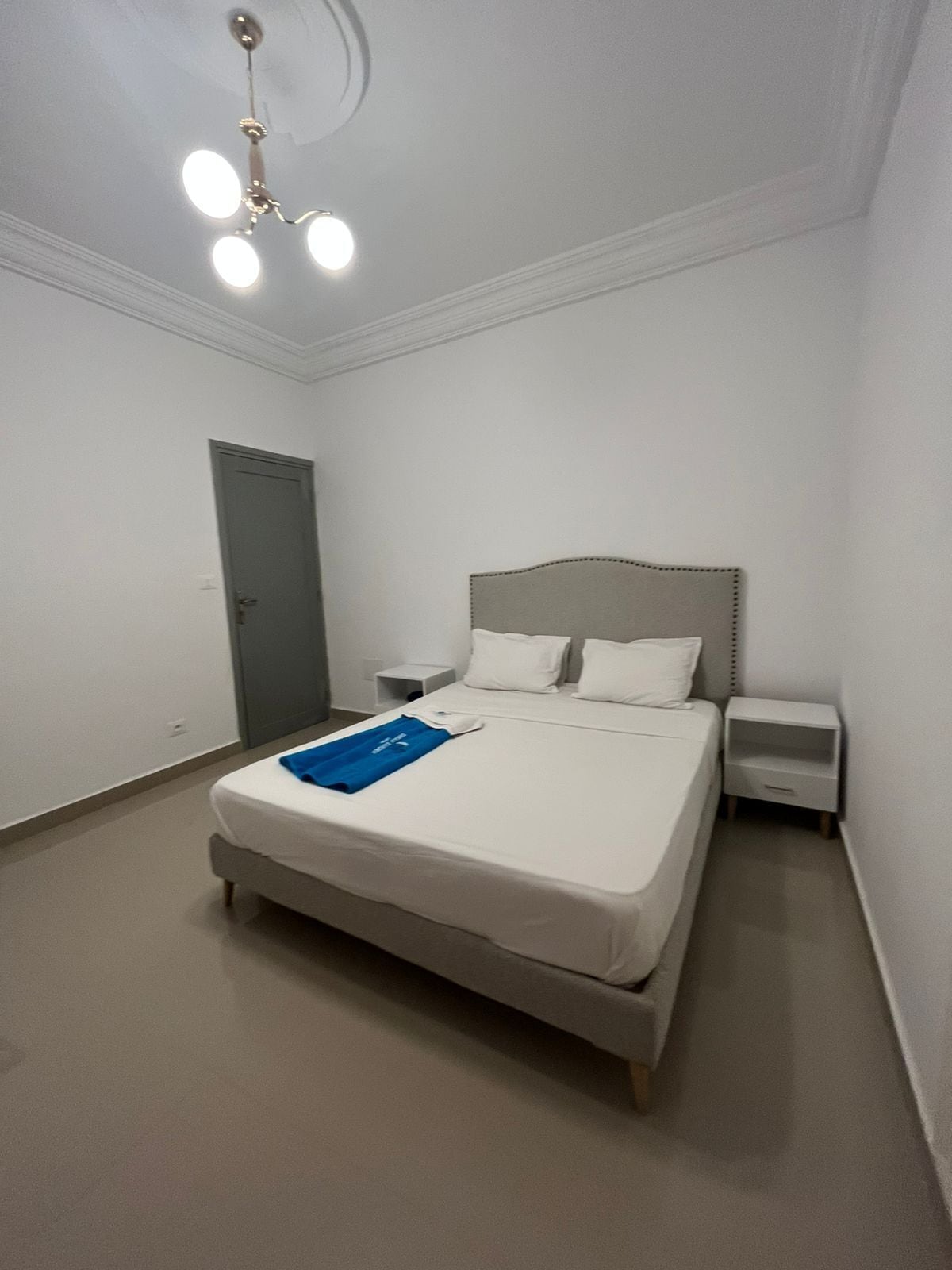 A bedroom with a bed, two bedside tables, a door, and a ceiling light fixture.