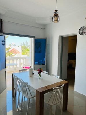 A dining area with a wooden table and white chairs, a vase with flowers, and a view of a balcony with a red-roofed building. - Dream Garden Djerba (Mezraia)