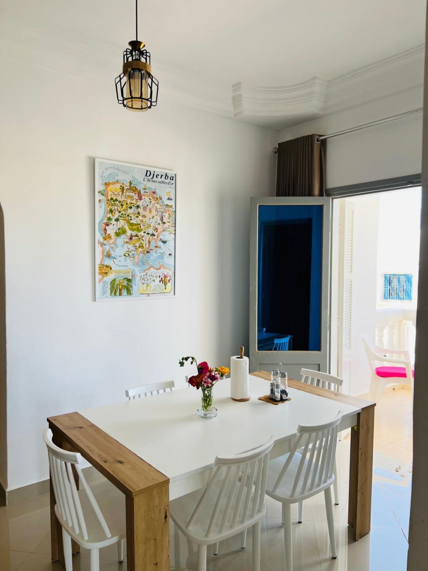 Un coin repas avec une table blanche, quatre chaises blanches, un banc en bois, un luminaire suspendu, une carte accrochée au mur et une vue sur l’extérieur.