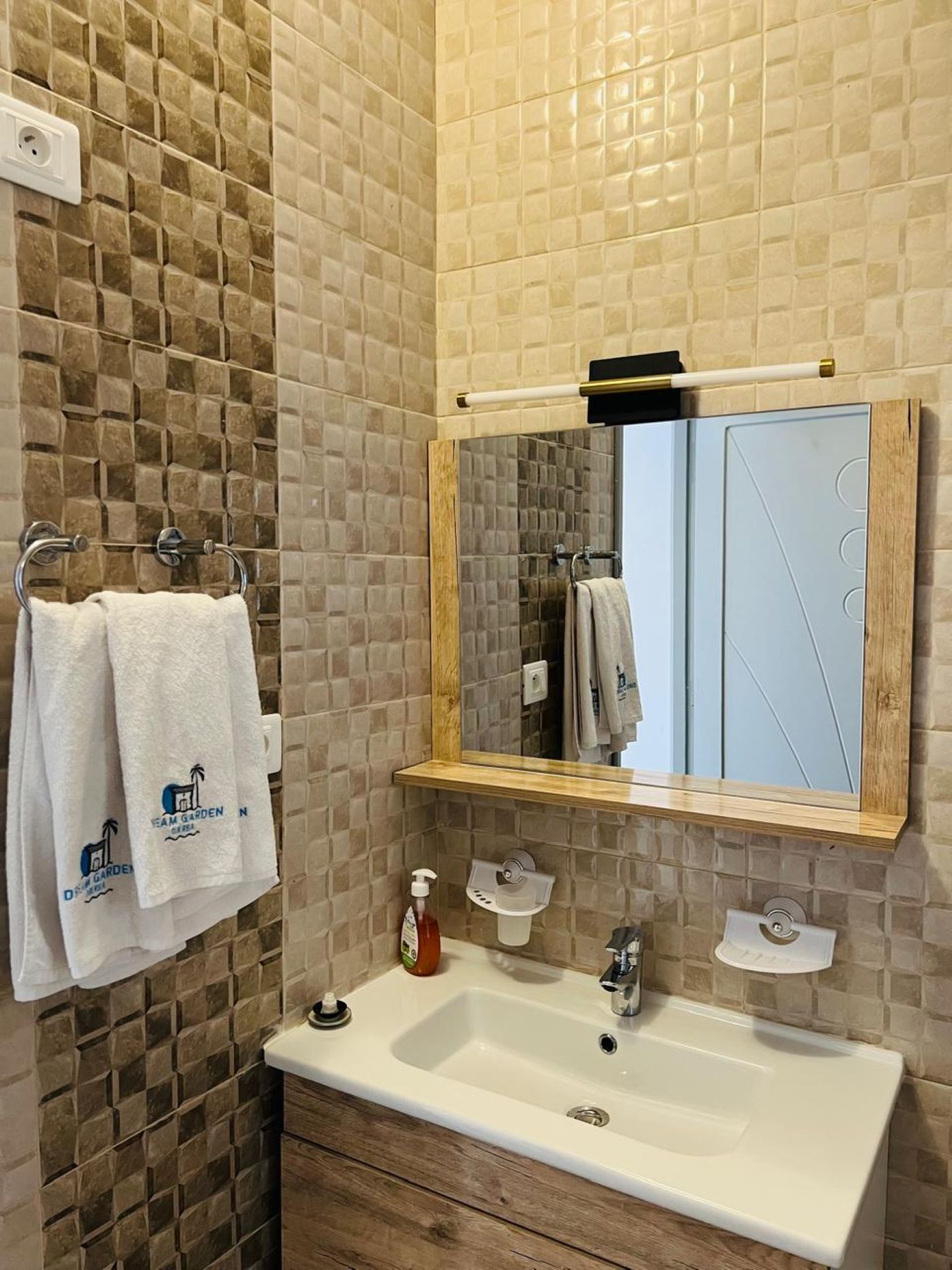 Un baño con pared de azulejos, tocador de madera, lavamanos, espejo y dos toallas colgadas en un perchero.