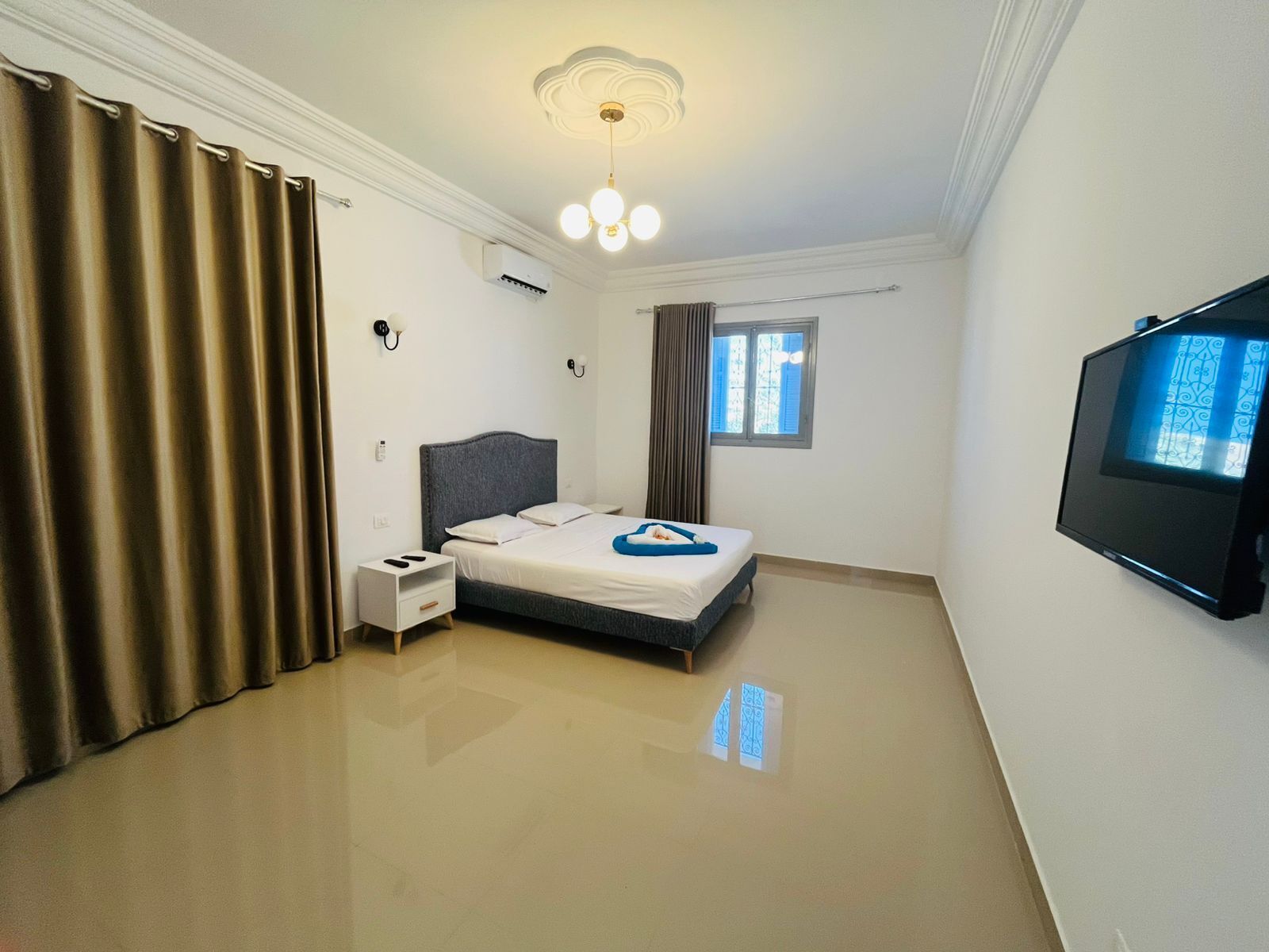 A bedroom with a bed, bedside table, curtains, a ceiling light, an air conditioning unit, a window, and a television.