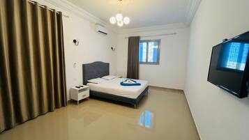 A bedroom with a bed, bedside table, curtains, a ceiling light, an air conditioning unit, a window, and a television.