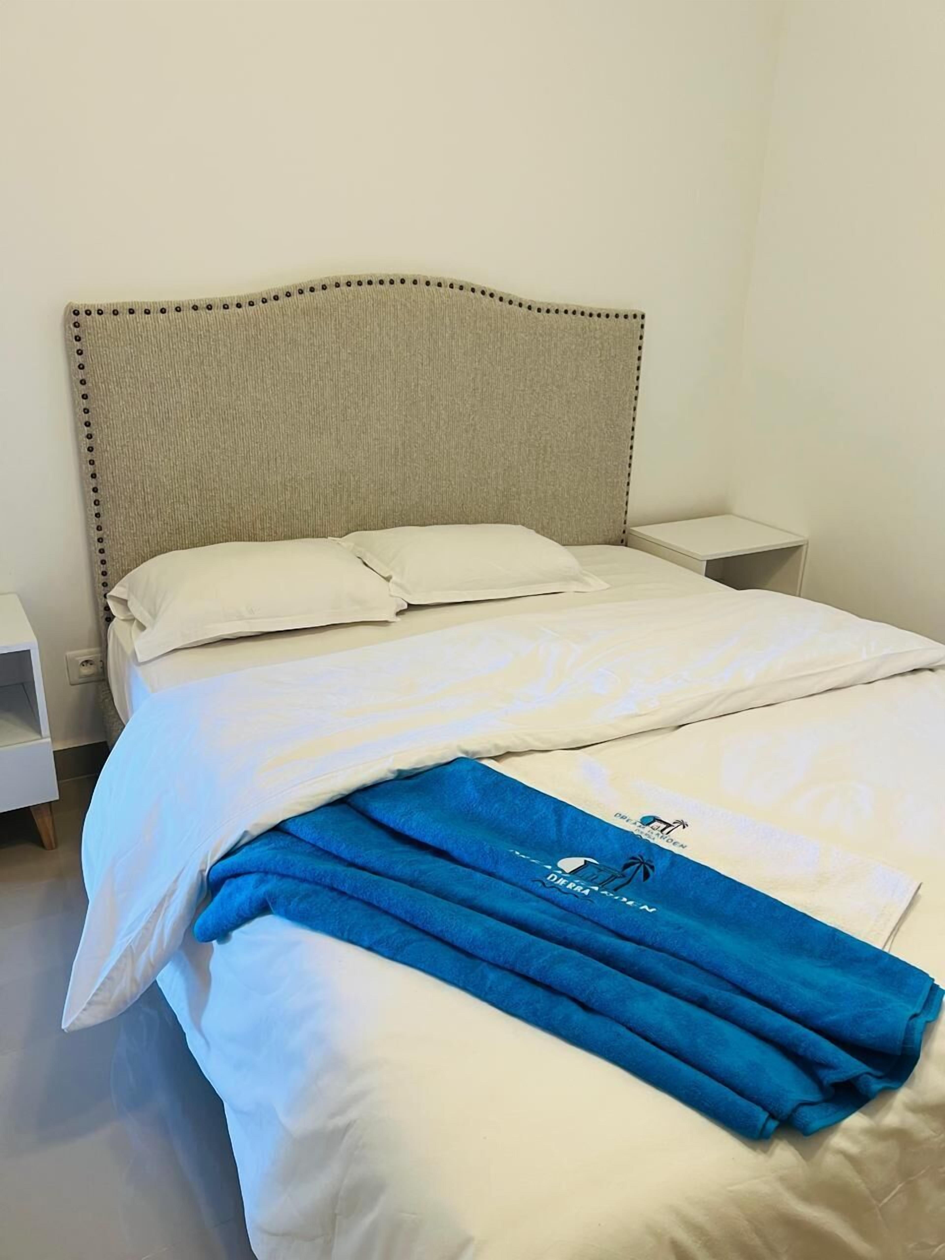 A neatly made bed with white linens and a blue towel, a plain headboard, and a small bedside table.