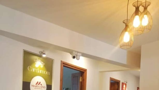 A hotel corridor with a poster advertising Cardamom Maryland, a wooden door, and a hanging light fixture. - Maryland Residency (Sulthan Bathery)