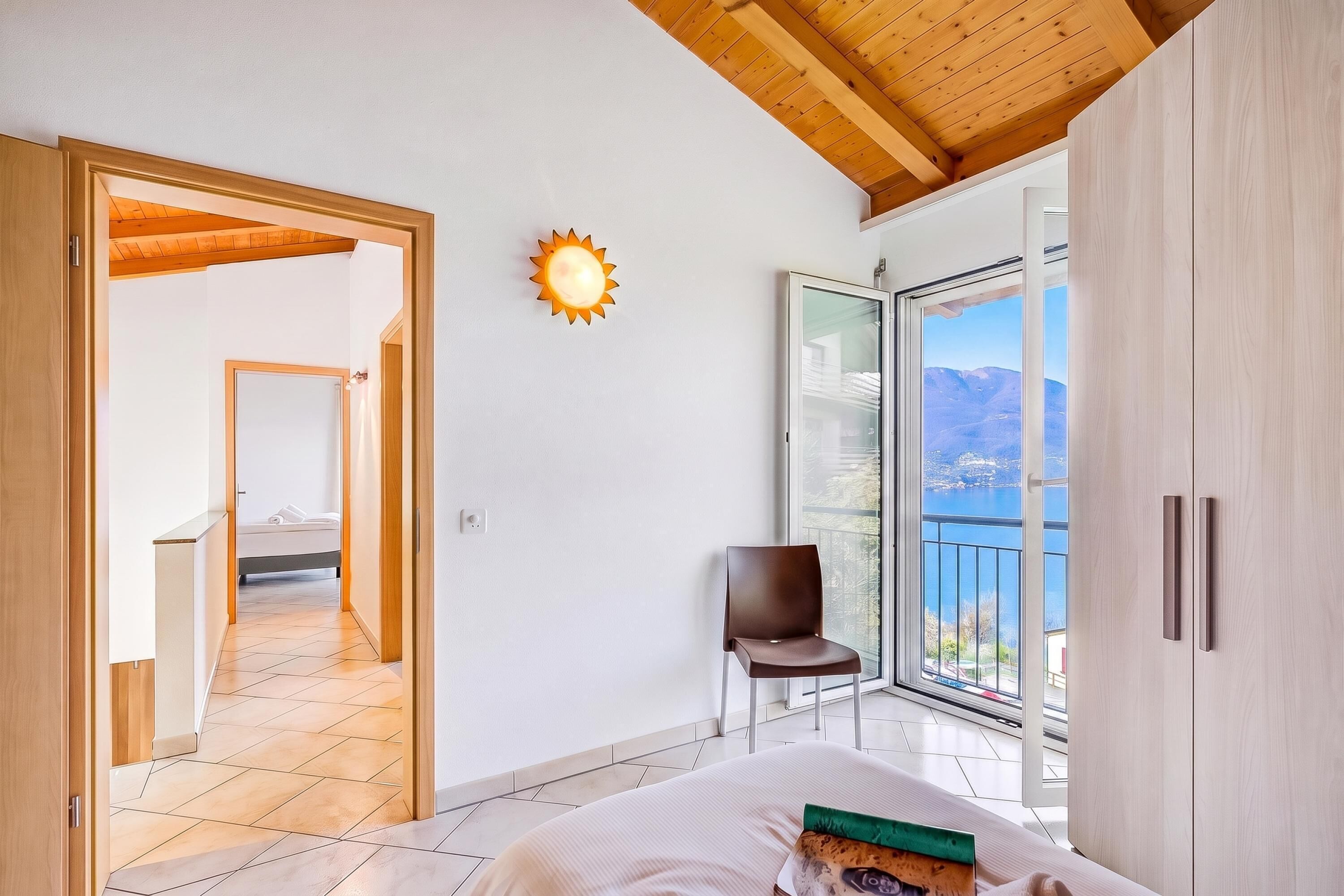 A bedroom with a bed, a chair, a sun-shaped light, and a balcony with a view.