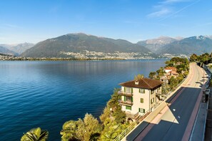 A lakeside hotel with a view of mountains, a paved road, and a clear sky. - Casa Zaira 1 (Gambarogno)