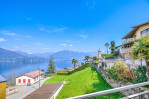 A lakeside view with a mountain in the background, a building with a clock tower, and a well-maintained lawn. - Casa Zaira 1 2BR in Caviano Gambarogno (Gambarogno)