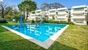 A swimming pool with a mountain backdrop, surrounded by a well-maintained lawn and a multi-story building with balconies. - Garden & Surf Residenza By Max With Pool & Balcony (Riva del Garda)