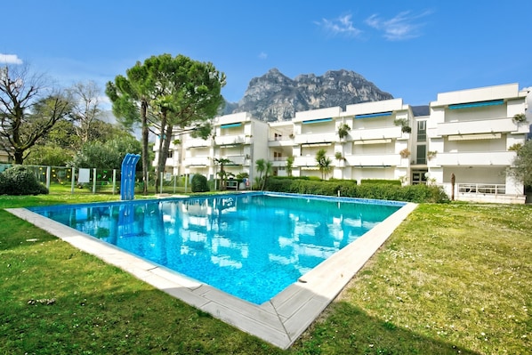 A swimming pool with a mountain backdrop, surrounded by a well-maintained lawn and a multi-story building with balconies.