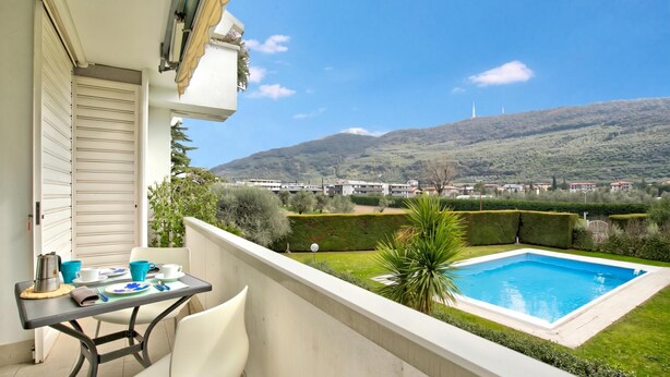 A balcony with a table, chairs, and a view of a swimming pool and distant buildings. - Garden & Surf Residenza By Max With Pool & Balcony (Riva del Garda)