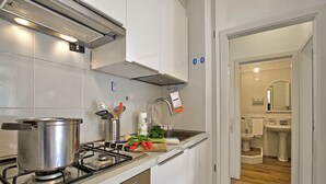 A modern kitchen with a gas stove, a pot, and various kitchen utensils. There is a view into the adjacent bathroom with a sink and toilet. - Garden & Surf Residenza By Max With Pool & Balcony (Riva del Garda)