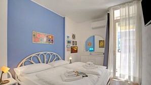 A neatly made bed with white linens and towels, a bedside lamp, and a wall with framed pictures and artwork. - Garden & Surf Residenza By Max With Pool & Balcony (Riva del Garda)