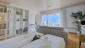 A hotel room with two beds, a sofa, a desk, a bookshelf, and a large window with a view. - Casa Miramonti Muralto With Garage and Garden (Muralto)
