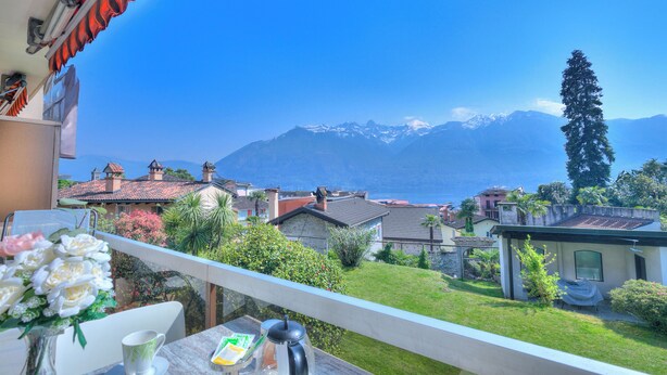 A balcony with a view of a mountain and lake, a table with a vase of flowers, and a chair. - Casa Miramonti Muralto With Garage and Garden (Muralto)