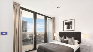 A modern hotel room with a large bed, a nightstand, a framed picture, and a view of the city through a sliding glass door. - One Bedroom Apartment City View (London)
