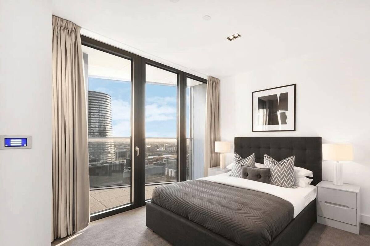 Une chambre d’hôtel moderne avec un grand lit, une table de chevet, un tableau encadré et une vue sur la ville, visible à travers une porte-fenêtre coulissante.