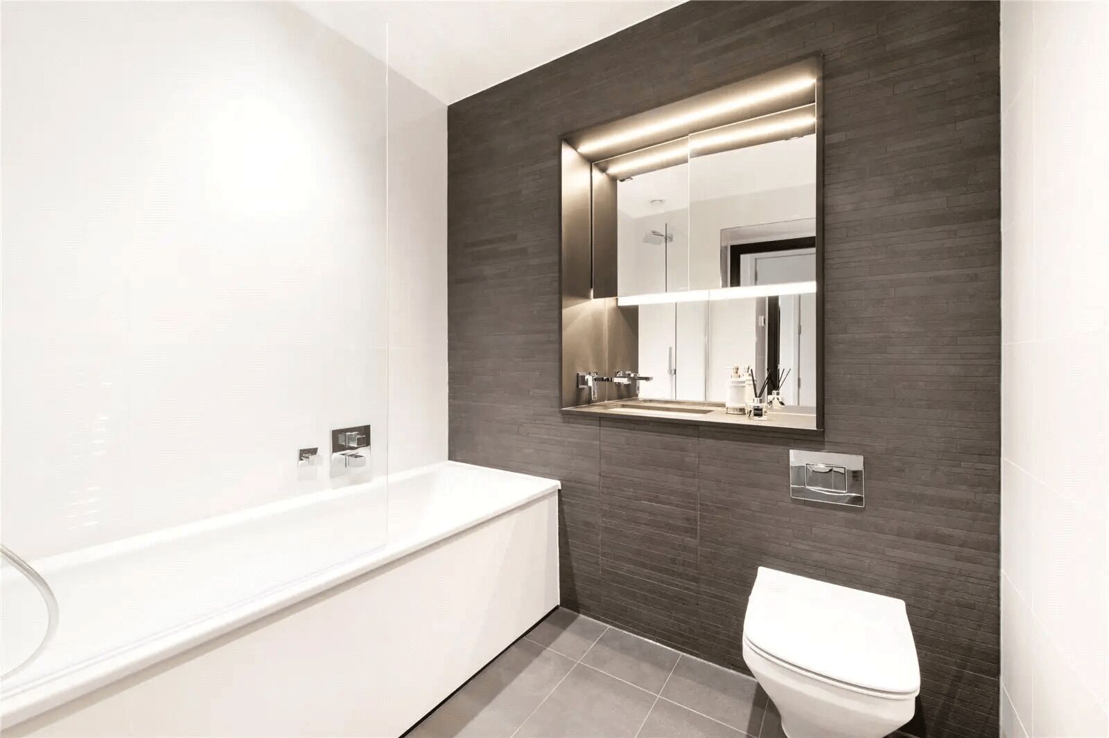 A modern bathroom with a white bathtub, a toilet, and a mirror with integrated lighting.