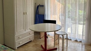 Une pièce comprenant un lit, une armoire blanche, une table ronde avec un pied rouge et une fenêtre ornée de rideaux légers.