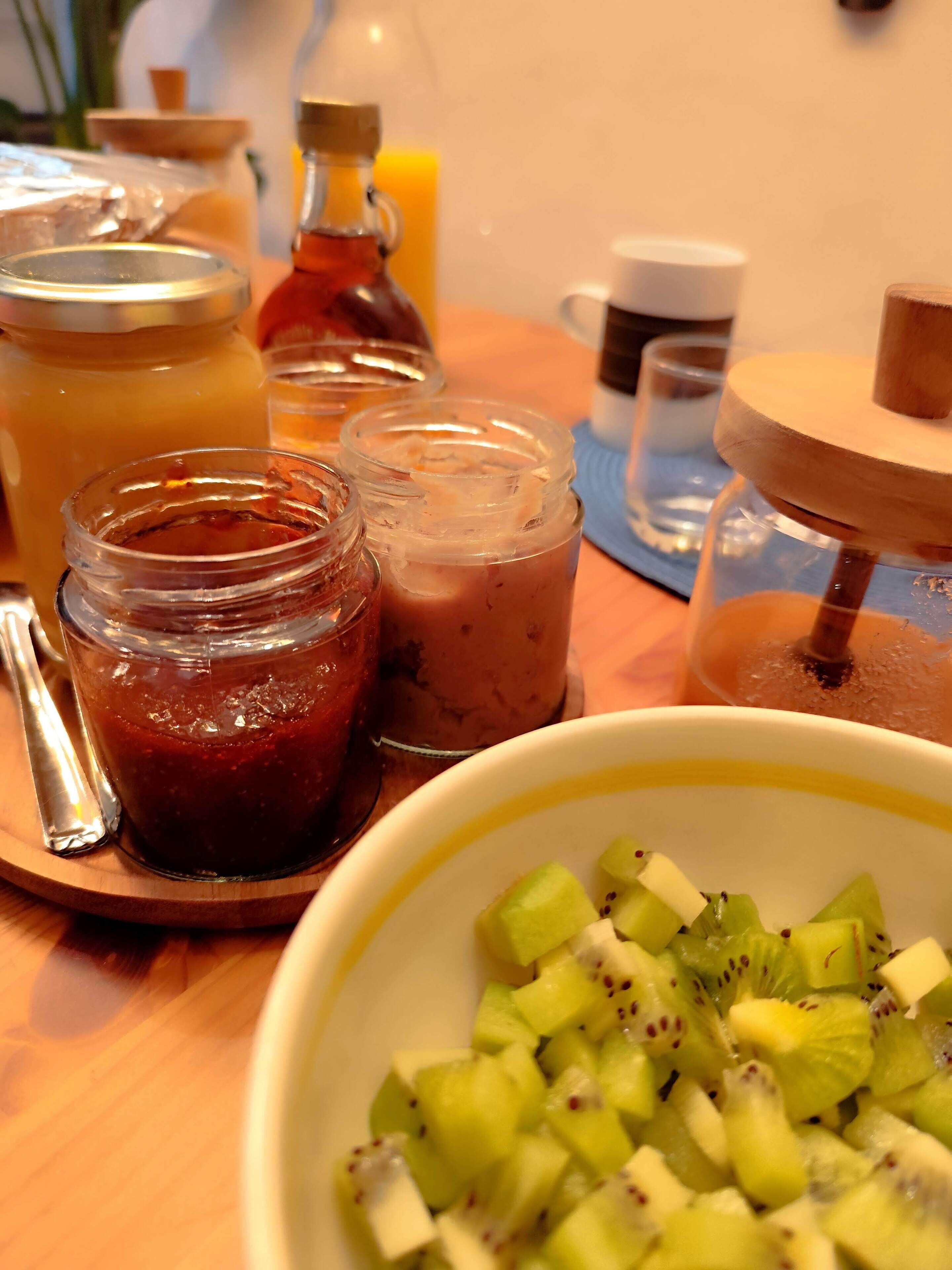 A wooden table with a bowl of sliced kiwi, a spoon, and several glass jars containing different food items.