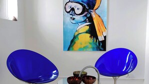 Deux chaises bleues, une table en verre placée entre elles, un tableau accroché au mur et une fenêtre donnant sur un paysage.