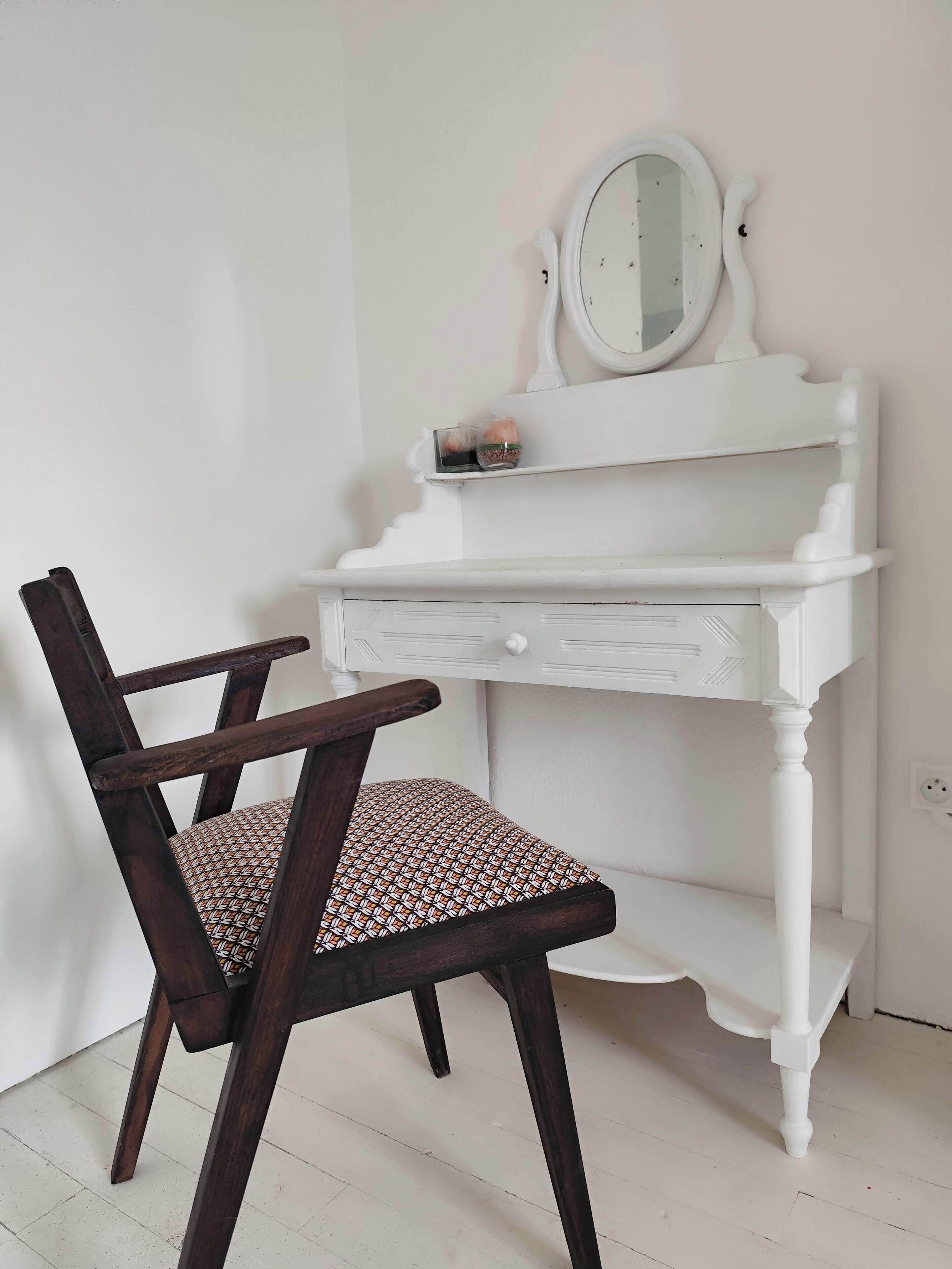 Une coiffeuse blanche avec un miroir et une petite étagère, une chaise en bois foncé avec un coussin à motifs, et un sol blanc.