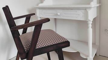 Une coiffeuse blanche avec un miroir et une petite étagère, une chaise en bois foncé avec un coussin à motifs, et un sol blanc.
