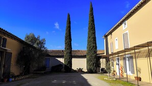 Une cour intérieure agrémentée de deux cyprès majestueux, d’un chemin de gravier et de bâtiments aux toits et fenêtres recouverts de tuiles.
