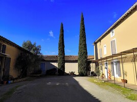 Un patio con dos altos cipreses, un camino de grava y edificios con tejados de tejas y ventanas.