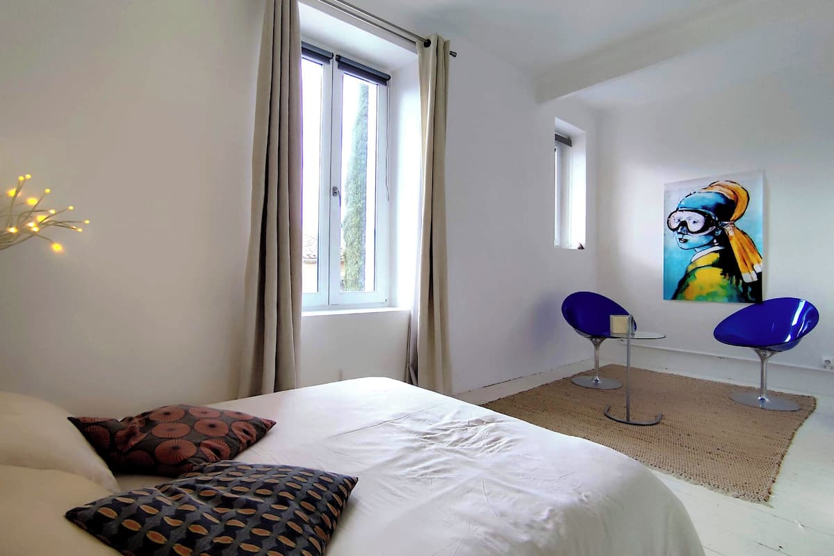 A bedroom with a bed, two chairs, a painting, and a window.